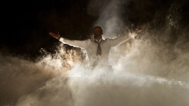 Backlit performer on stage with arms outstretched amidst lots of smoke