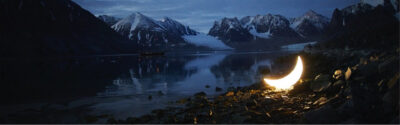 Electric light in the shape of a crescent mood placed on a pebble beach with snowy mountains in the background