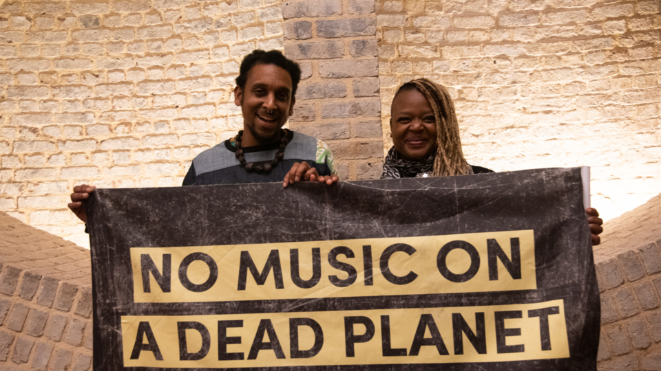 Ian and Eska stand holding a sign saying 'No Music on a Dead Planet