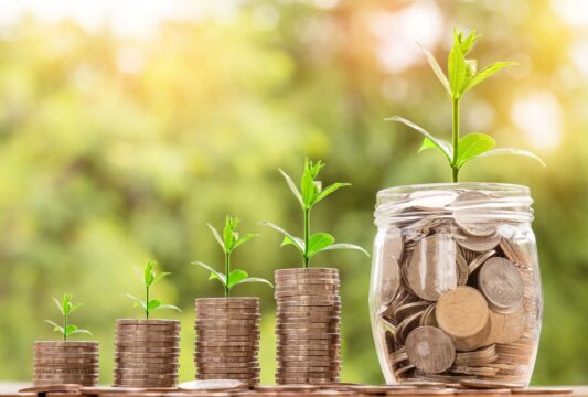 Four stacks of coins in ascending height, each with a small sprout then a jar of money with a larger sprout