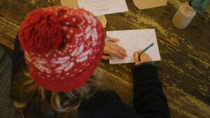 Close up from above and behind of someone with a red woolly hat writing a letter with right hand