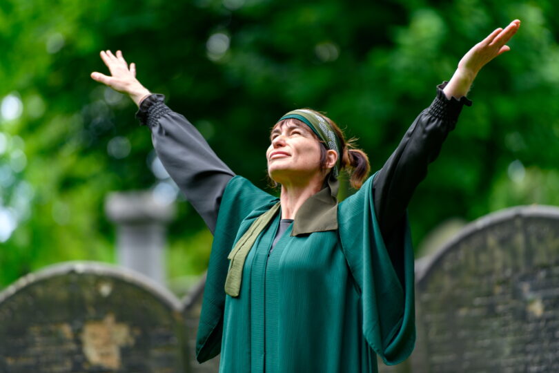 A woman is standing in a park with gravestones with her arms raised and looking at the sky