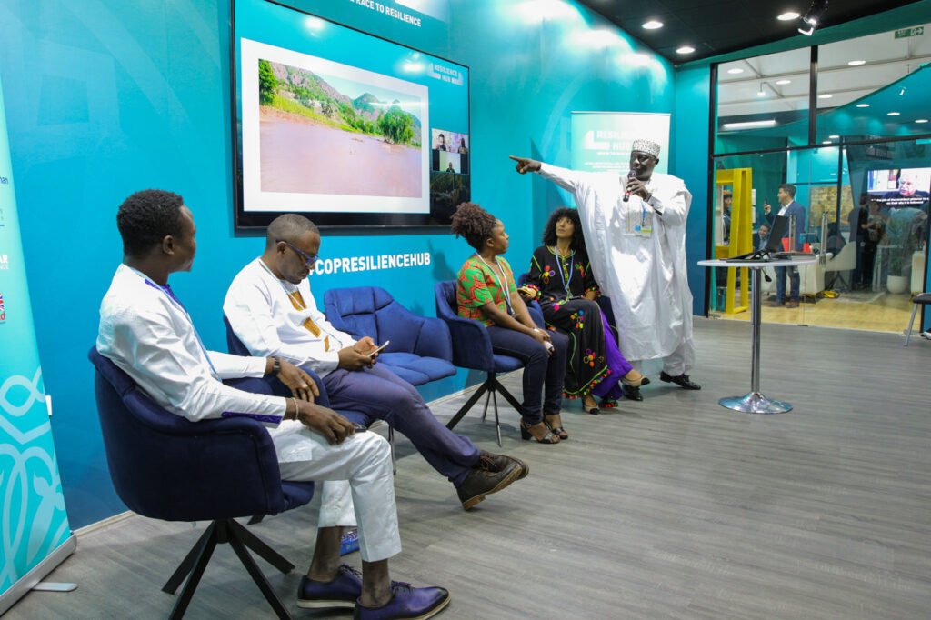 A row of people sitting on stage. A man wearing a kaftan is pointing to an image on screen whilst talking into the microphone