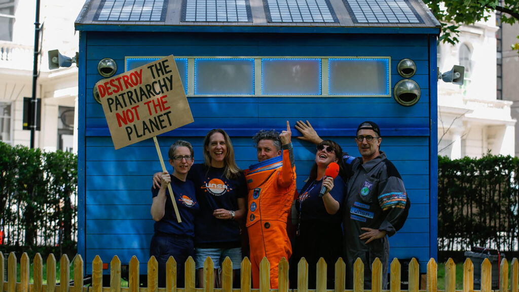 A group of smiling people with protest signs stand in front of a building 