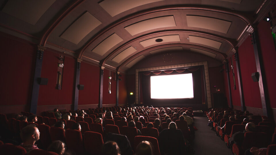 A seated crowd are watching a screen in a cinema. It looks like an old building with panelled walls and ceilings