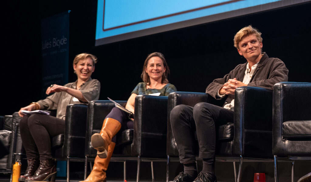 Three people sit on a stage as part of a panel discussion - they are smiling adn laughing.