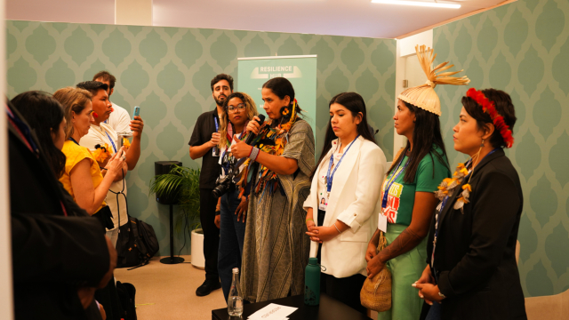 A group of people stand ing front of a crowd or spectators in a room. A woman in indigenous dress is addressing them. with a microphone