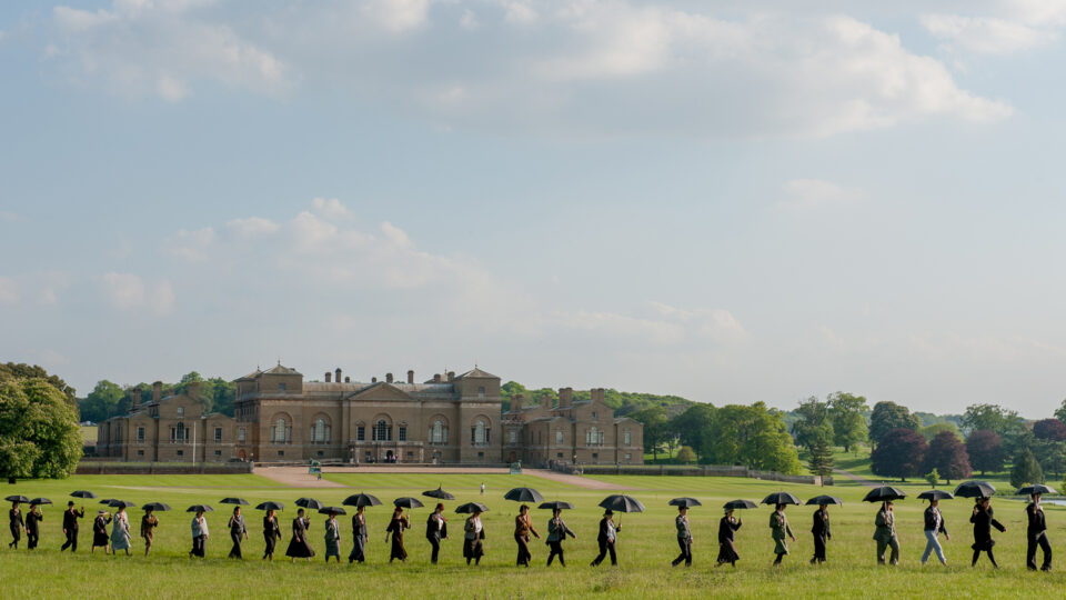 A row of people walking with black umbrellas on a sunny lawn, background is an old countryhouse-type building