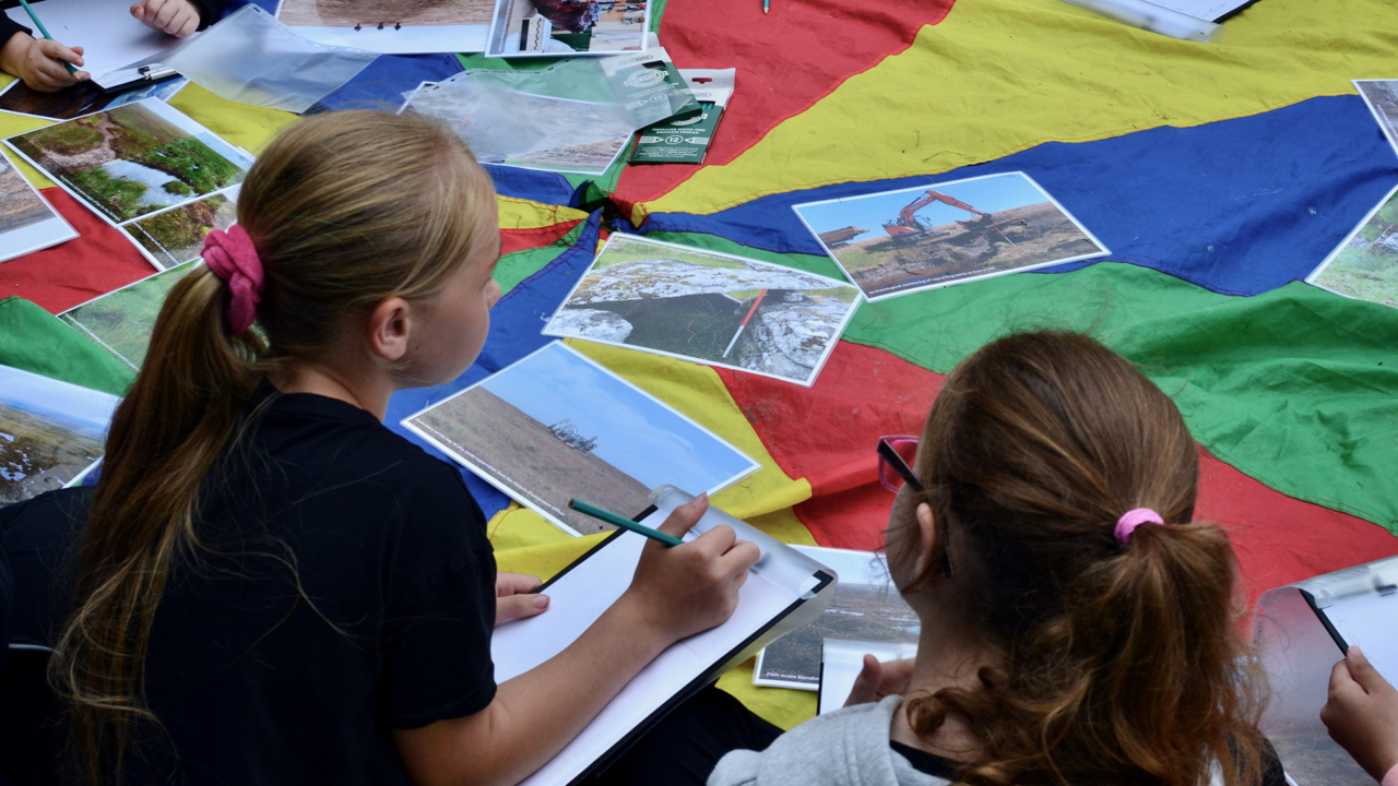 2 kids sat down on a table with lots of A4 photographs splayed across. They are in the process of writing.