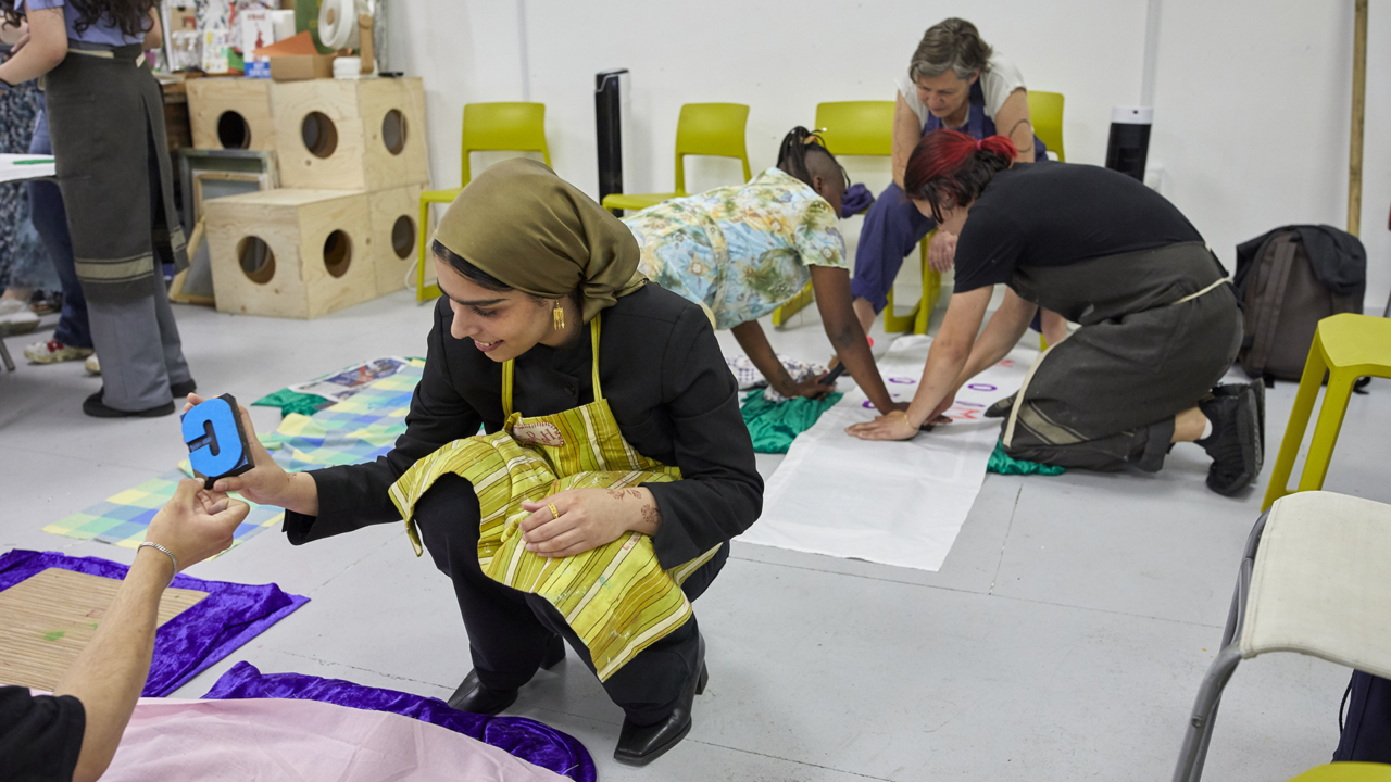 A woman is kneeling in a studio creating some art which is laid out on the floor. In the background a group are working together, also creating artwork on the floor.