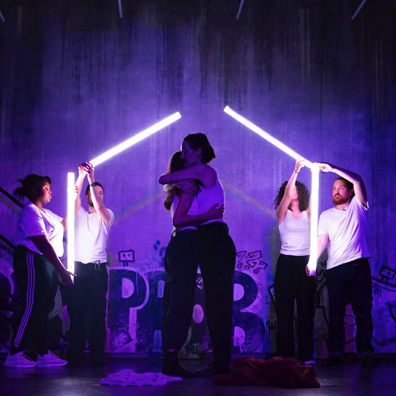 Theatre production on stage, purple lit stage with 4 people in the background holding up strobe lights, and two people in the foreground hugging.