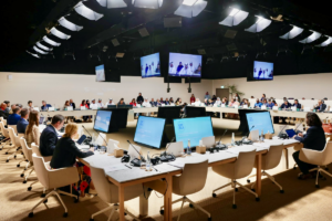 A room of government officials at a COP meeting. They sit in a square shape facing inwards. There are many monitors on desks in front of them and also hanging from the ceiling.