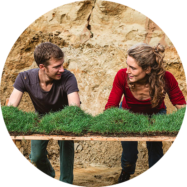 Two people slightly bending over and looking at eachother, there are patches of grass in front of them on a table. Background is a rocky wall.