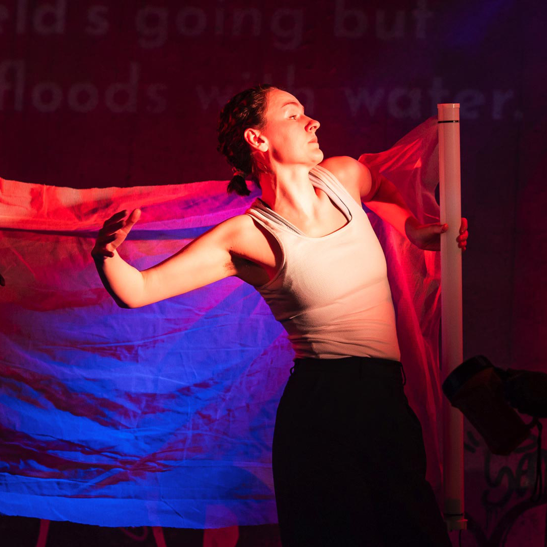 Spotlight shot of a person on the left performing with a white top and black trousers, and chiffon fabric wrapped around them. Silhouette of a person on the right looking on.