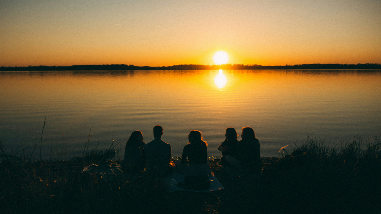 a group of people sitting on the shore of a lake at sunset