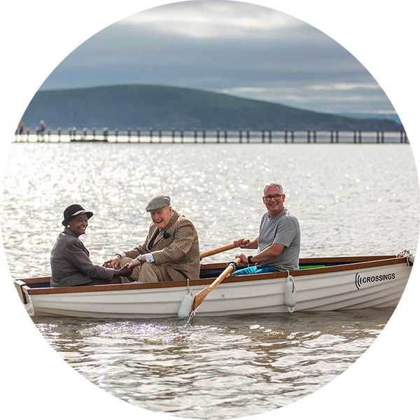 Three people on a rowboat, on a lake with hill in background, they are smiling and looking at the camera