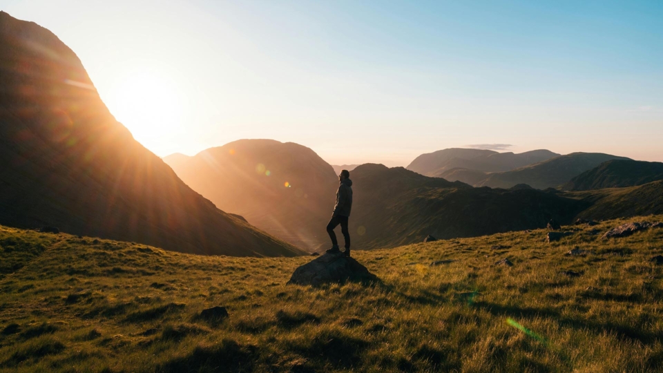 Silhouette Photography of Person Standing on Green Grass in Front of Mountains during Golden Hour