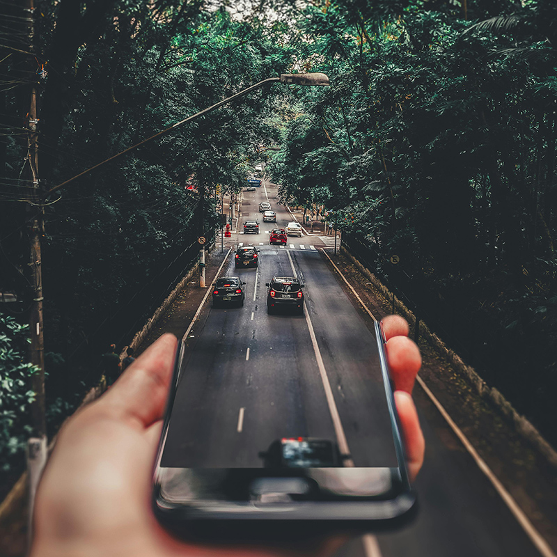 an abstract image of someone holding a smartphone and it merges to the background of a road with cars and lots of trees