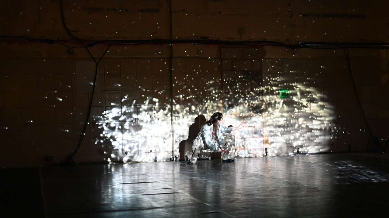 A dance performance in a dark room. The performer is lit up by a projection featuring scattered specks of light resembling a galaxy in space - she is kneeling on the floor and reaching out.